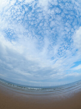 Strong fish-eye distortion emphasises a huge expanse of intricately clouded blue summer sky over a curved horizon of beach, sea and wind farm.