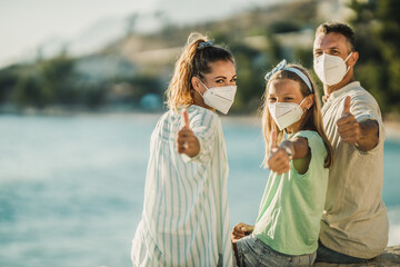 Family With Protective Mask Sitting Near The Sea With Thumbs Up And Looking At Camera
