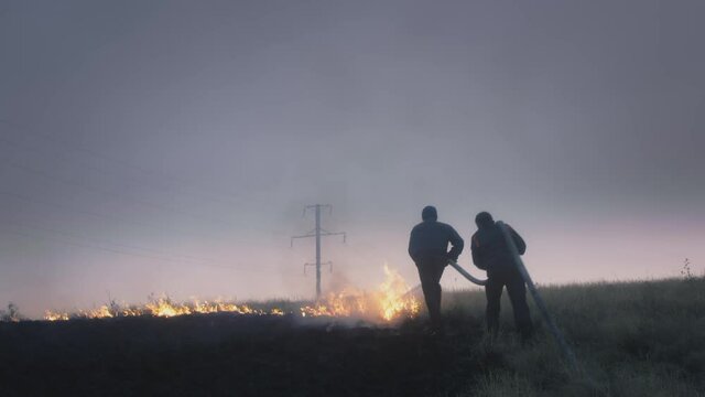 Real two firefighters carry a hose with water in their hands, extinguish the burning flame. Nature is on fire: grass, steppe and field. Water from a hose is sprayed onto the fire. Danger and smoke