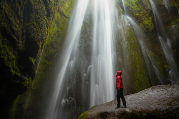 Woman wearing a red jacket standing on a wet boulder at the foot of a majestic waterfall in a...