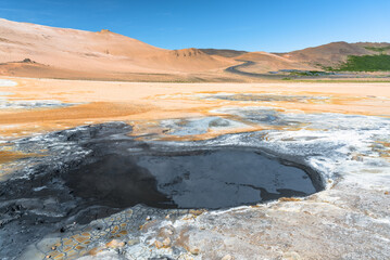 Boiling mud pool in a geothermal area in Iceland on a sunny summer day. A winding road  climbing a barren volcanic hill is visible in background. Hverir, Iceland.