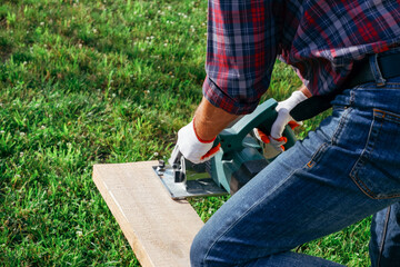 Working with a power tool. A builder is sawing a board at the construction site of a new house. Close-up on gloved hands holding a circular saw.