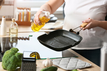 Mature woman pouring oil into frying pan in kitchen - obrazy, fototapety, plakaty