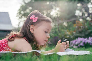 Portrait of little girl 3-4 in red lying on blanket on green grass and looking into mobile phone. Children using gadgets