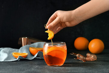 Woman decorating glass of tasty aperol spritz cocktail with orange on dark background, closeup