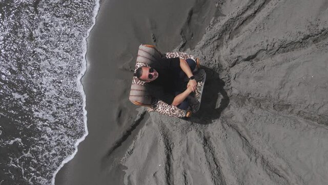 Guy with sunglasses is sitting in old chair in meditative and thoughtful pose, looking up, drone slowly spinning around, top view. Relaxing recreation after hard work or before important life changes.