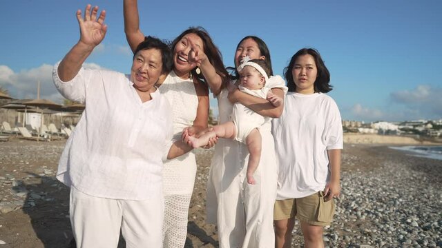 Group Of Multigenerational Asian Women And Baby Girl Waving In Slow Motion Smiling Standing On Picturesque Mediterranean Sea Coast. Happy Carefree Family Resting At Tourist Resort On Beach. Tourism