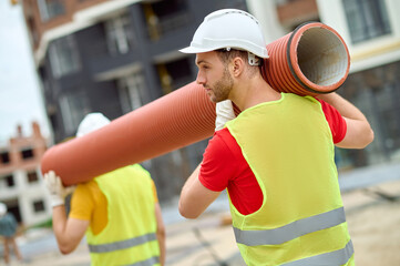 Two workers in protective helmets carrying a corrugated drain pipe