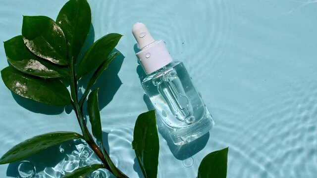 Glass cosmetic bottle with pipette lies on the surface of the water. Top view of water splash blue colored. Pure blue water with reflections sunlight. Sample of packaging for design, advertising.4k.