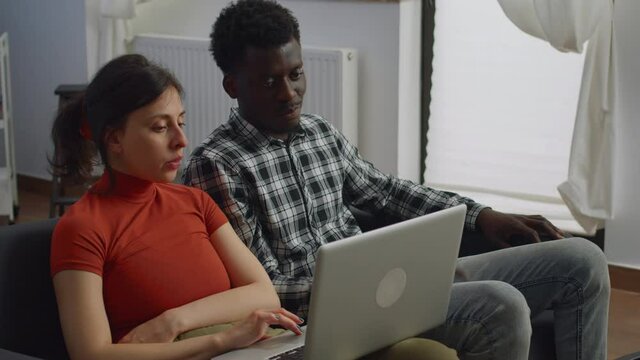 Interracial couple sitting on sofa using laptop in living room. Multi ethnic married people looking at screen together and talking. Mixed race husband and wife at home on sofa with technology