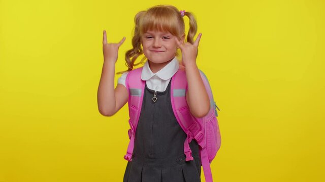 Overjoyed delighted funny pupil school girl teenage showing rock n roll gesture by hands, cool sign, shouting yeah with crazy expression, dancing, rejoicing in success on yellow studio background