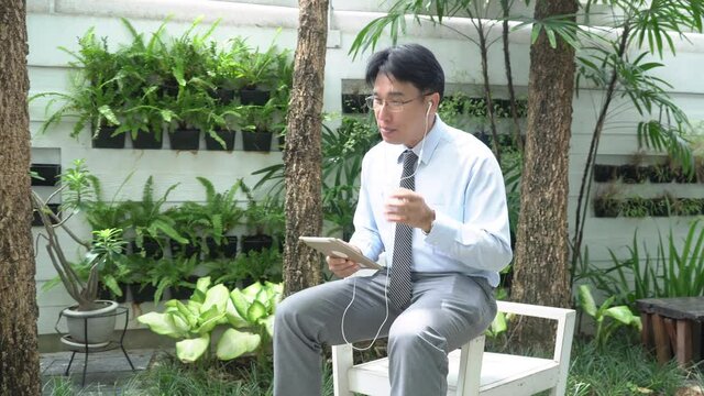 4K Asian businessman walking in the garden using digital tablet video conference business meeting discussion with colleague. Mature male working small business with using wireless technology at home.