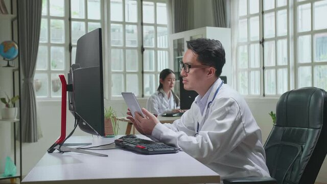 Asian Man Doctor Is Using Tablet While Work With Desktop Computer In Workplace. Medical Concept

