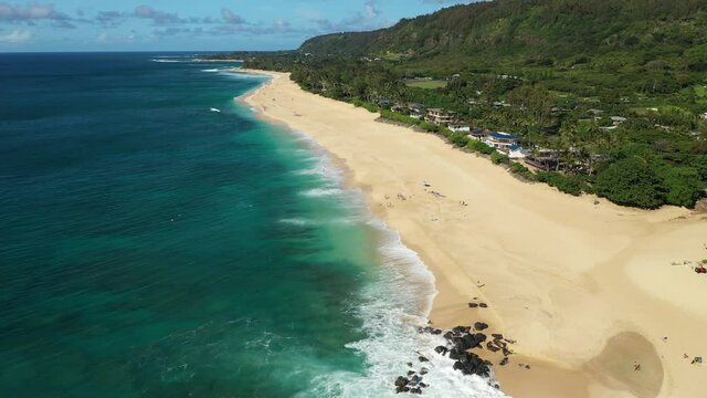 Aerial drone footage above a beach on Oahu, Hawaii, USA. Broad, sandy beach with turquoise water and white waves, and sunbathers visible on the sand. Green forest to the right of the beach.