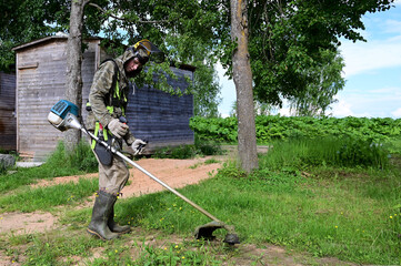 worker in special protective clothing with lawn mower in his hands mows grass. Trimer mows lawn. Lawn care with brushcutter mower or electric lawn mowing tool