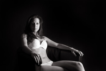 Elegant sexy woman sitting on an armchair, she is wearing white underwear in front of dark studio background, monochrome photo