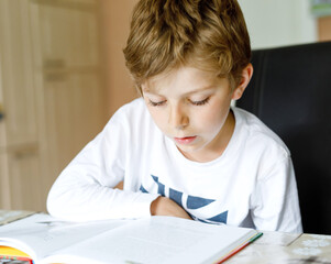 Naklejka na ściany i meble Little blonde school kid boy reading a book at home. Child interested in reading magazine for kids. Leisure for kids, building skills and education concept