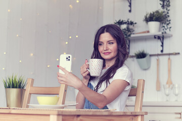 female smiling using mobile phone holding coffee at home in kitchen. ordering food through online stores. buying food through an online application