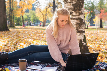 A young woman works in a park, typing on a laptop