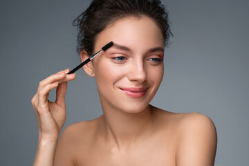 Young girl using brush for eyebrows. Photo of woman with perfect makeup on gray background. Beauty...