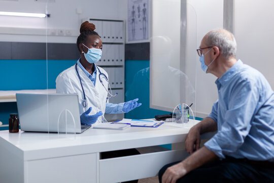 Doctor Of African American Ethnicity Wearing Face Mask While Talking To Elderly Patient At Desk In Medical Office. Black Medic And Old Man Discussing Healthcare Treatment During Epidemic