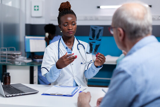 African ethnicity woman with doctor job looking at x ray scan in hand while discussing with senior ill patient at healthcare clinic. Black medic holding radiography for old man with disease