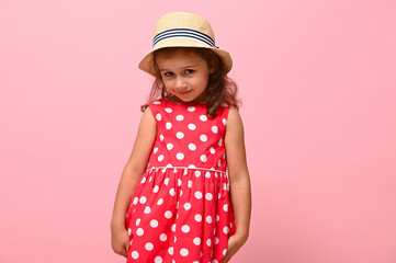 Pretty girl in a pink summer dress with white polka dots and a straw hat, looking shyly at the camera, posing against a pink wall background. Portrait of a happy smiling adorable child 3-4 years old.