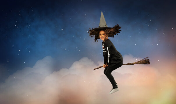 halloween, holiday and childhood concept - smiling african american girl in black costume dress and witch hat with broom over starry night sky and cloud background