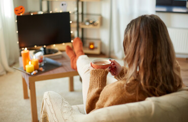 halloween, holidays and leisure concept - young woman watching tv and drinking hot chocolate with...