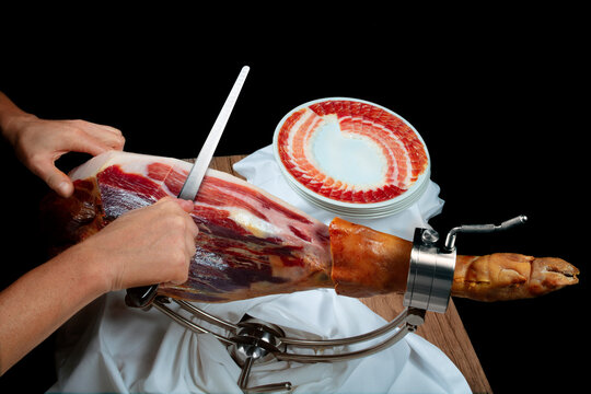 Mano cortando on cuchillo un ha on ib&eacute;rico y plato con jam&oacute;n sobre fondo negro. Hand cutting with knife an Iberian ham and plate with ham on black background.