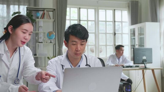 Hospital Medical Doctor'S Office: Two Asian Professional Physicians Talking, Consulting And Working On Laptop Computer. Specialists Discuss Patient Online Treatment, Writing Digital Medicine Prescript