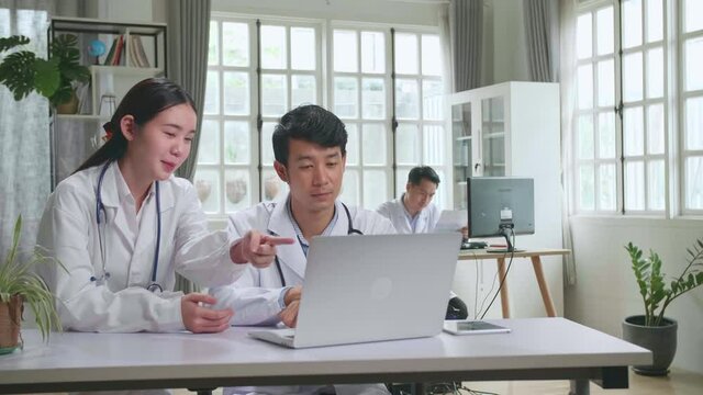 Hospital Medical Doctor'S Office: Two Asian Professional Physicians Talking, Consulting And Working On Laptop Computer. Specialists Discuss Patient Online Treatment, Writing Digital Medicine Prescript