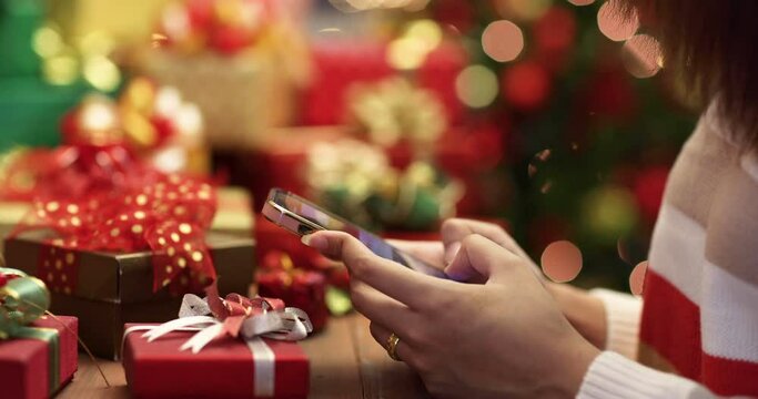 Unrecognizable woman in thick winter clothes sitting and desk with a pile of gift boxes and typing messages on smartphone screen with Christmas tree and bokeh lights in blur background.