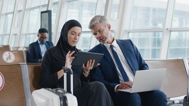 Caucasian Attractive Man And Muslim Woman In Business Style Sitting In The Hall Of The Airport With Suitcases, Speaking And Discussing Something At Their Gadgets