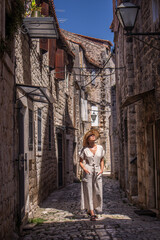 Fototapeta na wymiar Beautiful blonde young female traveler wearing straw sun hat sightseeing and enjoying summer vacation in an old traditional costal town at Adriatic cost, Croatia.