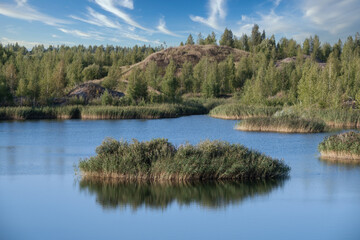 Fototapeta na wymiar A lake with a plane surface and reflections against the blue sky and white clouds. Originated from an old quarry flooded, groups of islands overgrown with reeds. Beauty in nature, landscapes, scenic.