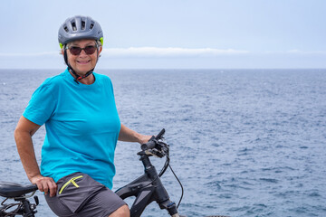 Happy mature cyclist woman enjoying healthy activity at sea with her electro bike. Standing on the cliff