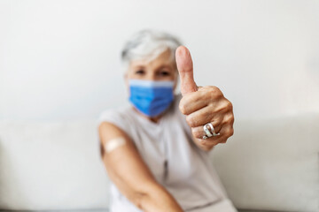 Woman with protective mask showing thumb up, and looking at camera, after she just got vaccinated. Portrait of senior women getting vaccinated with bandage. Senior woman showing plaster on her arm