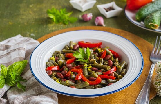 Warm salad or vegetable stew made from asparagus beans, canned red beans and roasted red bell peppers in a white plate on a green concrete background. Beans recipes. Vegan food.
