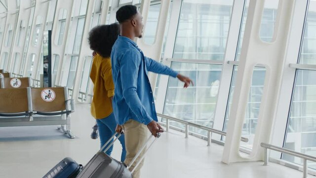 Multiracial family of three walking through the airport hall and looking at the window while carrying their suitcases on the wheels before the departure to the vacation