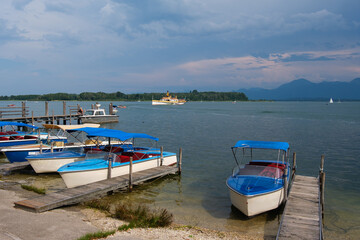 Fototapeta na wymiar Am Ufer vom Chiemsee in Bayern: Kleine Boote und ein Ausflugsschiff im Hintergrund - dahinter die Berge der Aplen