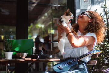 A young woman sits at a table at an outdoor cafe bar and plays with the cat.