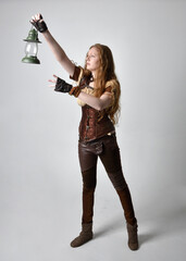 Full length  close up portrait of beautiful young woman with long red hair, wearing steampunk inspired costume holding  lantern  isolated on studio background.