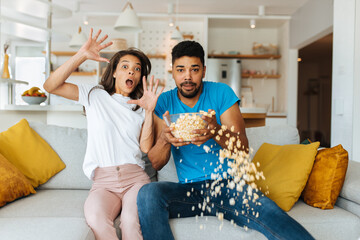 A surprised afro couple is sitting together at home and watching an action movie on television. A man is spilling popcorn.