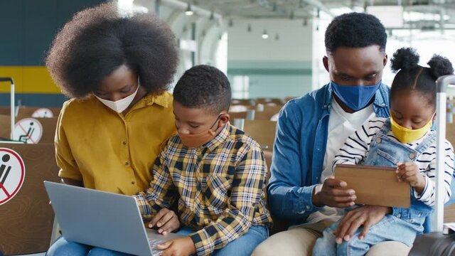 In standby. Waist up portrait view of the multiracial family wearing protective masks spending time with each other and watching something at their gadgets while waiting for their plane at the airport