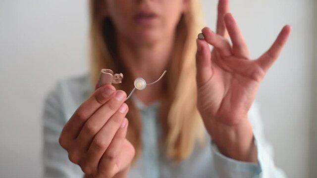 selective focus, blurred blond woman smile face  hold beige hearing aid in two hands in focus on light background. open devise and show how to use and put battery device for communication