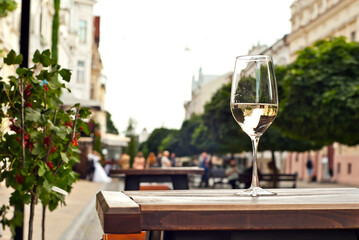 Glass of white wine close up. Alcoholic drink on a wooden table on a background of the city. Transparent wine on the summer terrace of the restaurant. Blurred people in the background.