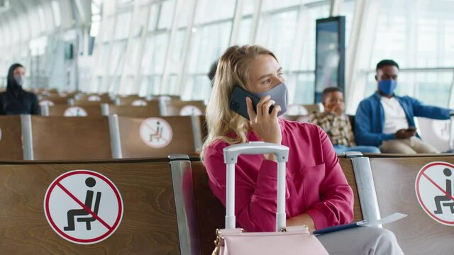 Caucasian gorgeous woman wearing protective mask sitting at the hall of airport with a smartphone in hand and answering the call while waiting for the departure