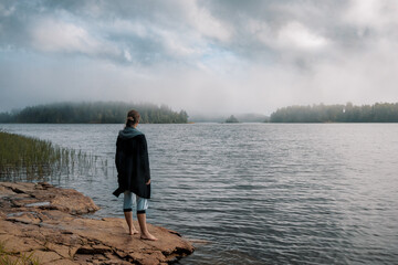 A girl with long hair in a blue jacket stands barefoot on a large stone on the shore of the lake and looks at the beautiful islands in the fog.