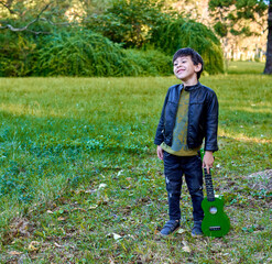 latino boy standing smiling in the forest holding a green ukulele at his side. square